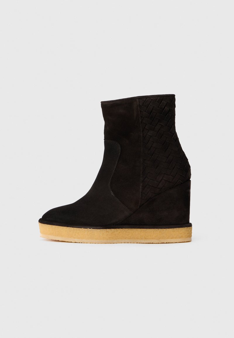 Bottines à talon compensé en daim noir avec détail tressé sur le côté et semelle en caoutchouc beige, présentées en profil latéral sur un fond blanc.