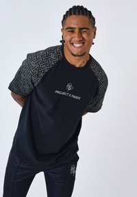 Camiseta negra con mangas cortas estampadas, que presenta un logo "PROJECT X PARIS". Combinada con pantalones oscuros, de tejido suave y corte casual.