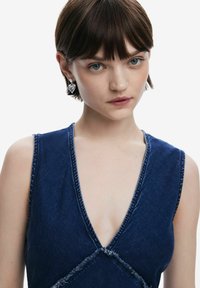 Mörkblå denimklänning med djup V-ringning, fransade kanter och subtila korsstygnsdetaljer. Bärs med ett hjärtformat silvörhänge.