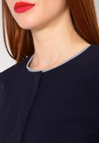 T-paita tummansinisellä värillä, jossa on pyöreä kaula-aukko vaaleanharmaalla reunuksella. Materiaalissa on suloinen tekstuuri ja kaulassa on huomaamaton napitus.