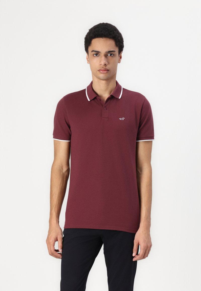 Polo shirt bordeaux avec un col classique, des bordures blanches et un petit logo brodé sur la poitrine gauche, fabriqué en un tissu en coton mélangé doux.