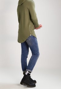 Sudadera con capucha de color verde olivo y dobladillo asimétrico, con detalle de cremallera lateral, combinada con jeans ajustados azul y botas negras de cordones.