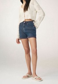 Dżinsowe szorty z wysokim stanem i podwiniętym brzegiem w ciemnym niebieskim kolorze, zestawione z krótkim, dzianinowym, białym swetrem o teksturowanym wzorze. Lekkie sandały.