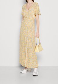 Robe maxi jaune à fleurs avec des manches courtes évasées, décolleté en V, design enveloppant, accompagnée d'un petit sac à main texturé jaune et de baskets blanches.