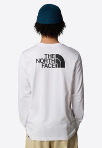 Witte lange mouwen shirt met een groot zwart "The North Face" logo op de achterkant. Gekoeld met een blauwe gebreide beanie. Eenvoudig ontwerp, zachte stof.
