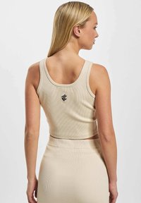 Beige ribbet crop top med brede stropper og rund halsudskæring, med et lille sort logo på ryggen. Glat tekstur; tætsiddende design.