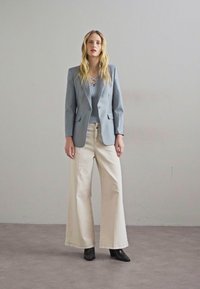 Jasnoszary blazer wykonany z gładkiego materiału, zestawiony z jasnoniebieską bluzką z detalem sznurowania oraz szerokimi spodniami w kolorze kremowym. Czarne botki do kostek.