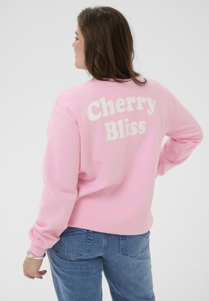 Kobieta stojąca z jedną ręką na biodrze, ubrana w jasnoróżową bluzę z napisem "Cherry Bliss" i niebieskie dżinsy na jednolitym tle.