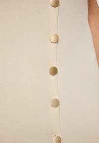 Rochie tricotată bej, cu o serie de nasturi aurii pe lateral, care evidențiază un material texturat și o siluetă slim.