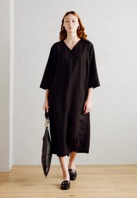 Robe noire midi avec un décolleté en V, des manches trois-quarts et des fentes latérales, associée à un grand sac noir et des chaussures noires à enfiler.
