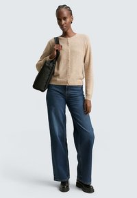 Béžový kardigán so zapínaním na gombíky, kombinovaný so širokými tmavomodrými džínsami. Na jednom ramene visí čierna kabelka. Hladká látka, voľný strih.