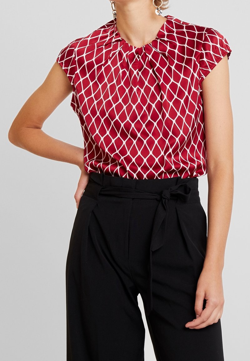 Femme portant un chemisier rouge sans manches avec un motif losanges blanc et un pantalon noir taille haute noué avec une ceinture en tissu, debout, main sur la hanche.