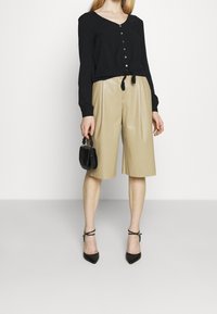 Blusa nera a maniche lunghe con bottoni e un fiocco in vita, abbinata a pantaloni corti plissettati beige e tacchi neri con cinturino alla caviglia, mentre tiene una piccola borsa nera.