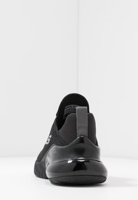 Czarne sportowe buty z siateczkową cholewką, teksturowanym wzorem i błyszczącem obcasem. Widok z tyłu pokazuje minimalistyczne logo i pętelkę do zakupu.