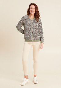 Bomberjacke mit floralem Muster und grünem Besatz, mit Reißverschluss und gerippten Bündchen, kombiniert mit hellbeigen Skinny Pants und weißen Sneakers.