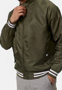 Veste bomber vert olive en nylon lisse, dotée de poignets rayés blancs et d'une fermeture éclair à l'avant. Comprend des poches latérales avec fermetures à pression.