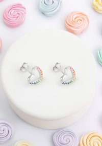 Sölvörhängen i form av hjärtan med flerfärgade stenar längs kanten, placerade mot en vit tårta med pastellfärgade svängningar.