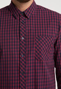 Czerwona i granatowa koszula w kratę z guzikami, z kołnierzykiem, kieszonką z przodu i ciemnymi guzikami. Materiał wydaje się gładki i ma wyważony krój.