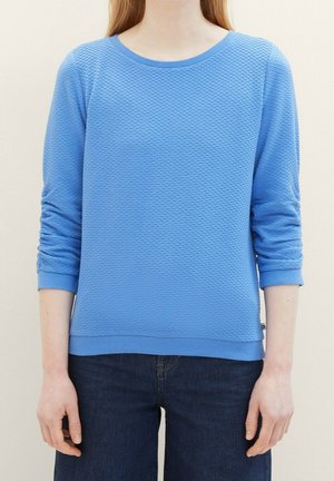 Person trägt ein strukturiertes hellblaues Langarmhemd mit gerafften Ärmeln und dunkle blaue Jeans, steht vor einem einfarbigen Hintergrund.