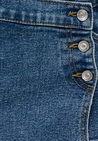 Jean en denim bleu devant avec trois boutons argentés visibles et des surpiqûres marron détaillées près de la taille et de la fermeture de la braguette.
