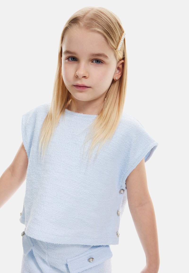 Hellblau strukturierte Bluse mit kurzen Ärmeln, versehen mit dekorativen silbernen Knöpfen an den Seiten. Das Model hat glattes, blondes Haar.