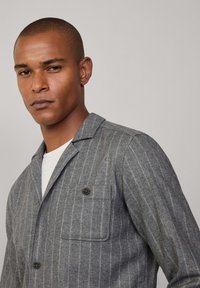 Giacca a righe grigia, tessuto testurizzato, taschino sul petto con bottone, collo a risvolto, indossata su una camicia bianca, design casual.