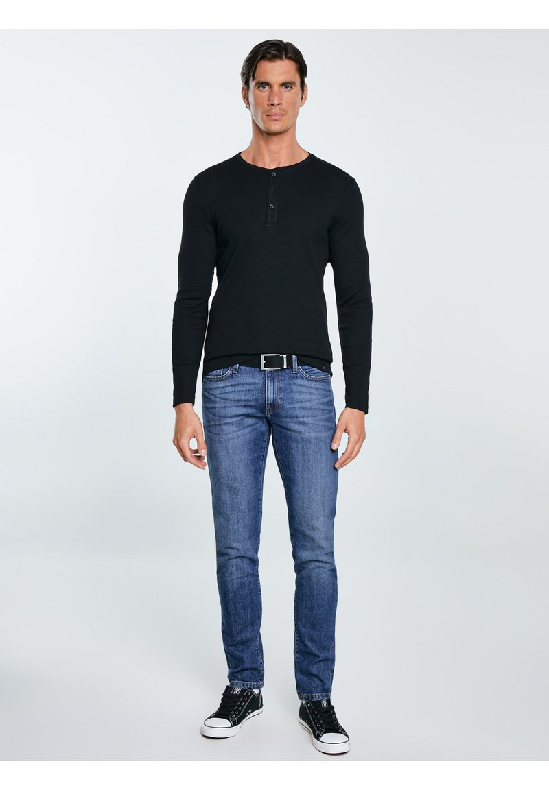 Mężczyzna stojący, ubrany w czarną, długorękawową koszulkę henley, niebieskie dżinsy, czarny pasek i czarne trampki z białymi czubkami, na neutralnym tle.