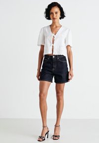 Bela kratka bluza s kratkimi rokavi in detajli iz čipke, v kombinaciji s temnimi denim kratkimi hlačami in črnimi sandali s peto ter belimi detajli.