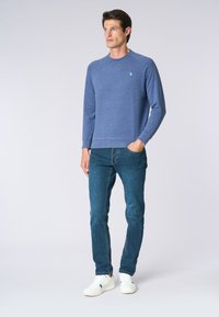 Felpa blu a maniche lunghe con scollo a girocollo e logo ricamato, abbinata a jeans blu attillati e sneaker bianche.