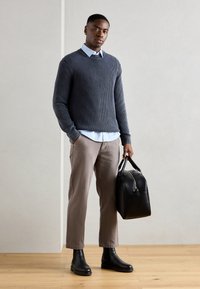 Mørk grå ribbestrikket genser over en lys blå skjorte med krage, kombinert med beige bukser og svarte skinn ankelstøvler, holder en svart veske.