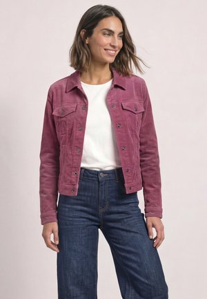 Femme portant une veste en velours côtelé rose boutonnée sur une chemise blanche avec un jean bleu foncé, souriante et regardant sur le côté.