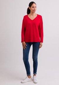 Roter V-Ausschnitt-Pullover aus weichem Material, mit langen Ärmeln und lockerer Passform, kombiniert mit schmalen blauen Jeans und weißen Turnschuhen.