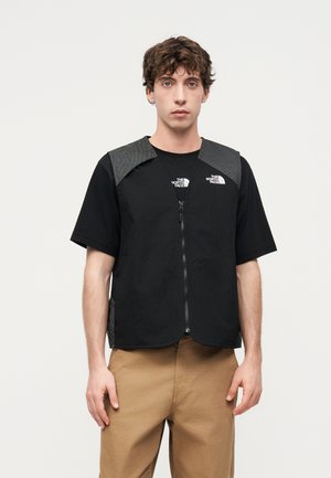 Young man wearing a black zip-up vest over a black short-sleeve t-shirt with The North Face logos and tan pants, standing against a white background.