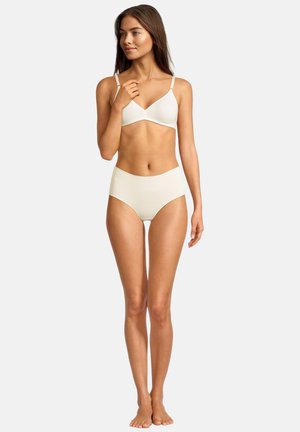 Junge Frau steht barfuß, trägt einen weißen nahtlosen BH und hoch taillierte Slips, schaut leicht lächelnd nach links.