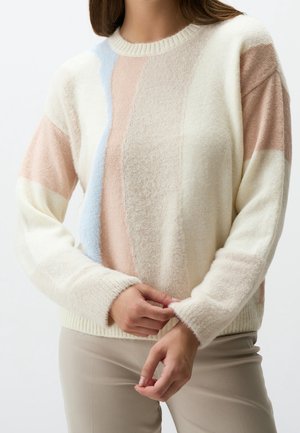Femme portant un pull doux texturé avec des blocs de couleur verticaux en crème, beige, rose clair et bleu pâle, ajustant le poignet de la manche.