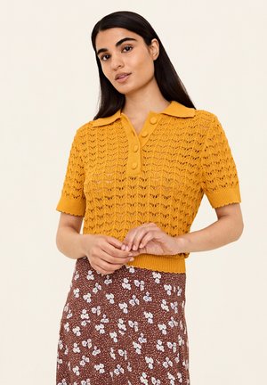 Maglione a polo a maglia color giallo senape con un motivo testurizzato, maniche corte e dettagli con bottoni sul colletto, abbinato a una gonna floreale marrone.
