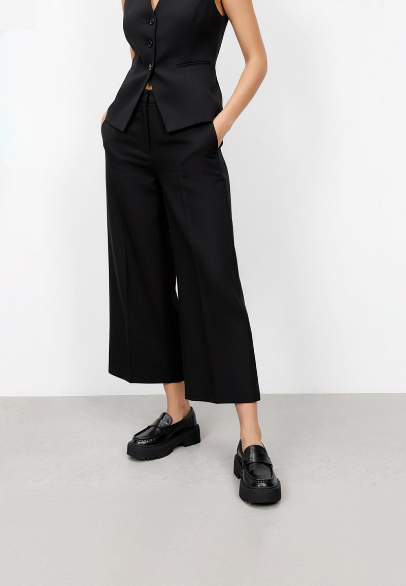 Gilet nero su misura con tre bottoni, abbinato a pantaloni neri a gamba larga. Mocassini neri lucidi con suola piatta completano l'outfit.