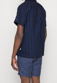 La camicia a maniche corte presenta strisce verticali nere e blu, un colletto e un orlo dritto. Abbinate a pantaloni corti azzurri.