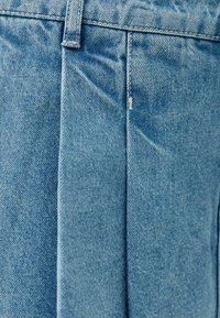 Tela de mezclilla azul claro con una textura suave, pliegues verticales y costuras resistentes en la cinturilla para un detalle adicional.