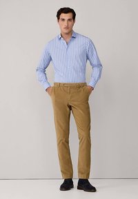 Niebieska w jasne paski koszula na guziki w połączeniu z beżowymi spodniami sztruksowymi. Model ma na sobie ciemne buty, ręce w kieszeniach, stoi na neutralnym tle.