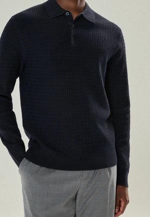 Homme portant un polo en tricot texturé noir à manches longues avec des boutons et un pantalon gris clair, les mains dans les poches, fond neutre.