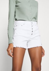Shorts de mezclilla blancos con bajo deshilachado, cierre de cinco botones y herrajes de cobre, combinados con un top verde de manga larga con botones.