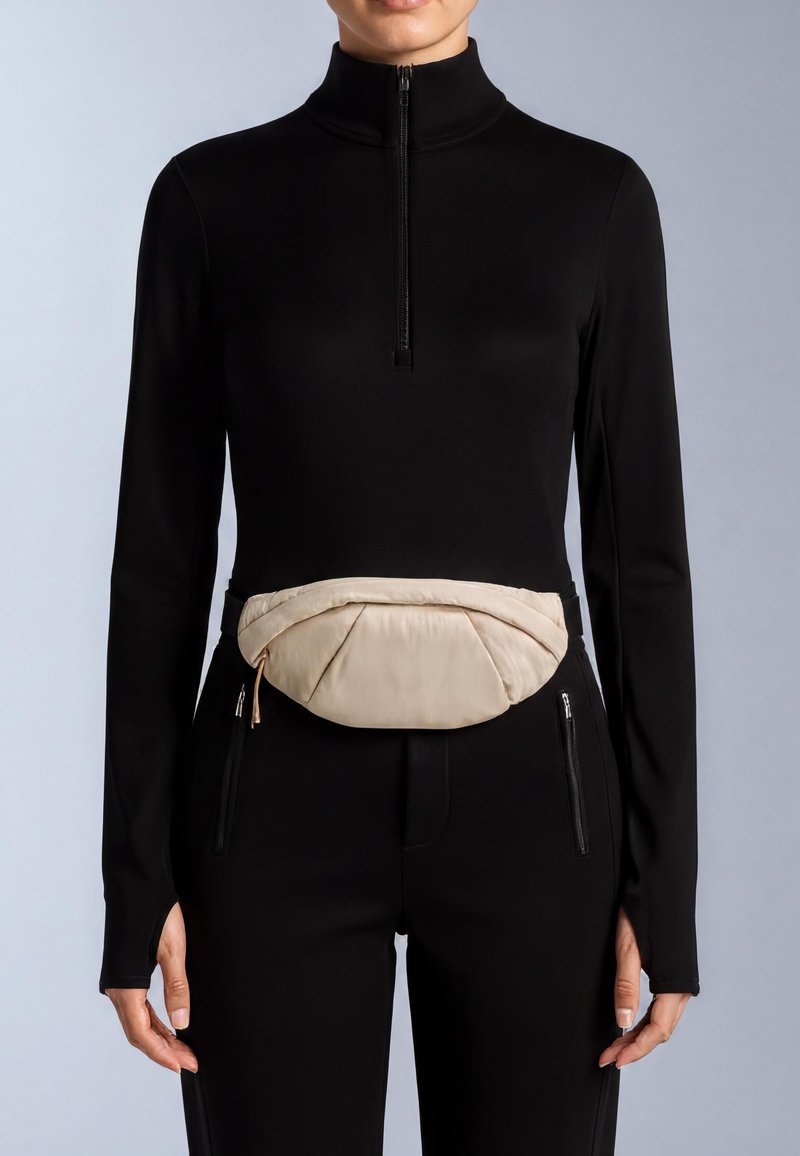 Beige tygbältesväska med en mjuk, vadderad design, bärs över en figurnära svart topp. Har blixtlås på sidorna, strömlinjeformad form.