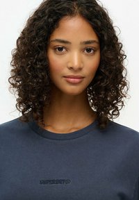 Granatowa koszulka z krótkim rękawem, wykonana z gładkiego materiału, z delikatnym, haftowanym logo "SUPERDRY" na klatce piersiowej. Okrągły dekolt.