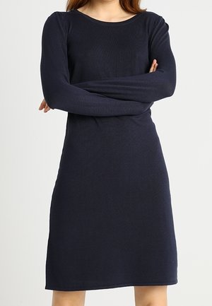 Kobieta ubrana w granatową sukienkę z długim rękawem sięgającą kolan, stojąca z założonymi skrzyżowanymi rękami na jasnym, jednolitym tle.