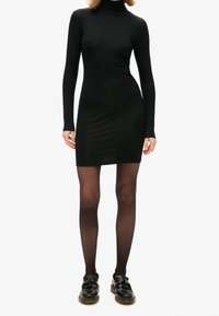 Frau in einem enganliegenden schwarzen Langarm-Turtleneck-Minikleid, transparenten schwarzen Strumpfhosen und schwarzen Quasten-Lackschuhen, stehend vor einem weißen Hintergrund.