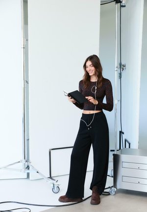 Musta pitkähihaine paita, jossa on hienovarainen kuvio, löysät mustat housut ja ruskeat kengät; pitelee muistikirjaa, johon on liitetty kuulokkeet.