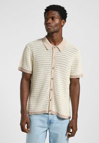 Kortärmad stickad polo i off-white med ett strukturerat mönster, beige krage och knappslå, med en avslappnad passform och ribbade kanter.