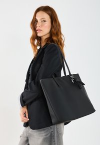 Sac fourre-tout noir en matériau lisse, doté d'une fermeture sécurisée, de longues poignées et d'un charme décoratif. Forme rectangulaire, design minimaliste.