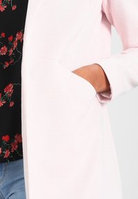 Light pink coat with a textured fabric, featuring a concealed pocket and a minimalistic design. Accented by a floral-patterned top underneath.